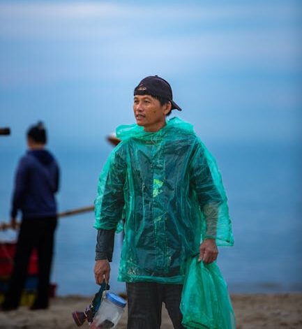 A man on the beach wearing a green rain poncho, holding gear, with a calm sea backdrop.
