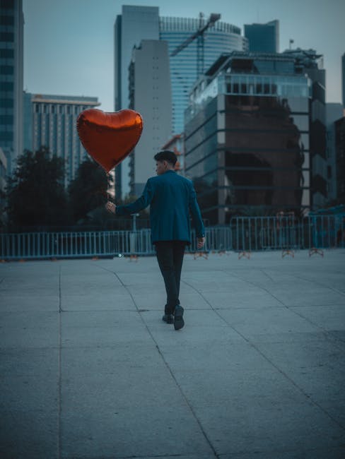 A man holding a red heart-shaped balloon walks through a city setting, capturing a romantic urban vibe.
